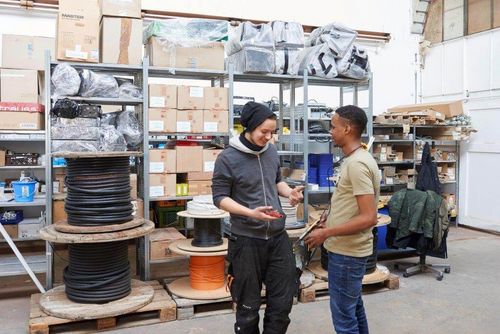 Zwei Männer stehen in einem Lagerraum und unterhalten sich. Im Hintergrund sind Regale mit Kartons, Kabelrollen und Materialien. Titel: Gespräch im Lagerraum