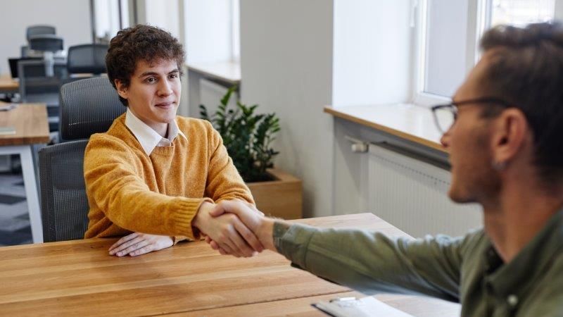 Ein junger Mann im gelben Pullover reicht einem Personaler über den Schreibtisch hinweg die Hand