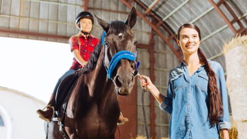 Eine Frau führt ein braunes Pferd am Halfter durch einen Stall. Auf dem Pferd sitzt ein ca 8-jähriges Mädchen. Beide lachen.