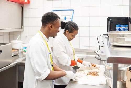Zwei Köche schneiden Fleisch in einer Profiküche; sie tragen weiße Jacken und Handschuhe.