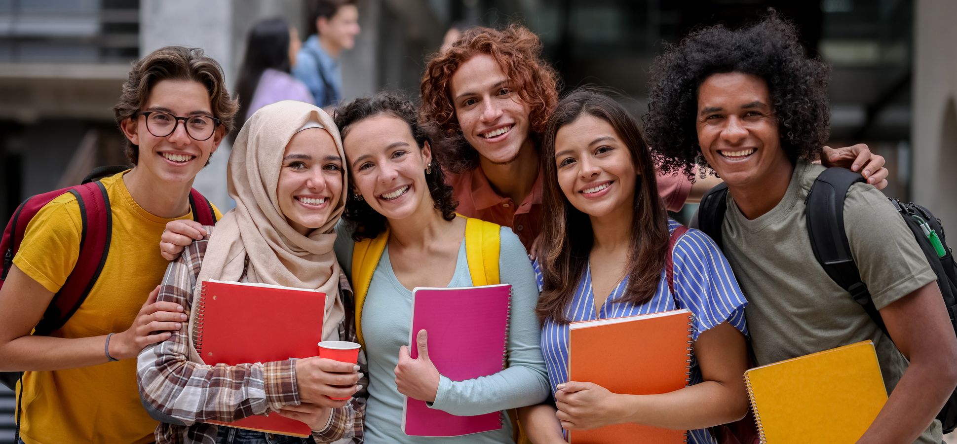 Sechs stehende Studierende mit Notizbüchern auf dem Campus.