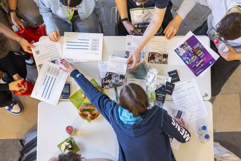 Aufnahme von oben auf einen Messetisch. Flyer und Infomaterial ist ausgelegt. Eine Unternehmensvertreterin erklärt den anwesenden Schülerinnen die Ausbildungsmöglichkeiten.