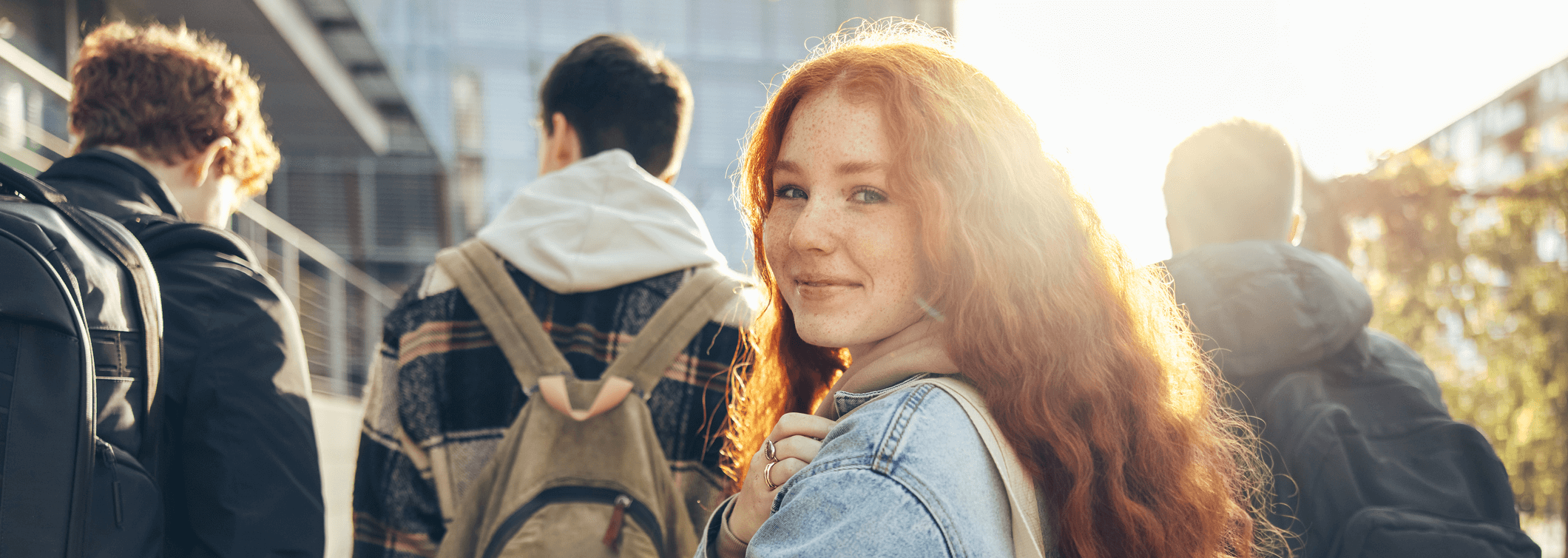 Teenager mit roten Haaren blickt über die Schulter in die Kamera. Im Hintergrund sind drei Jugendliche zu sehen, mit dem Rücken zur Kamera.