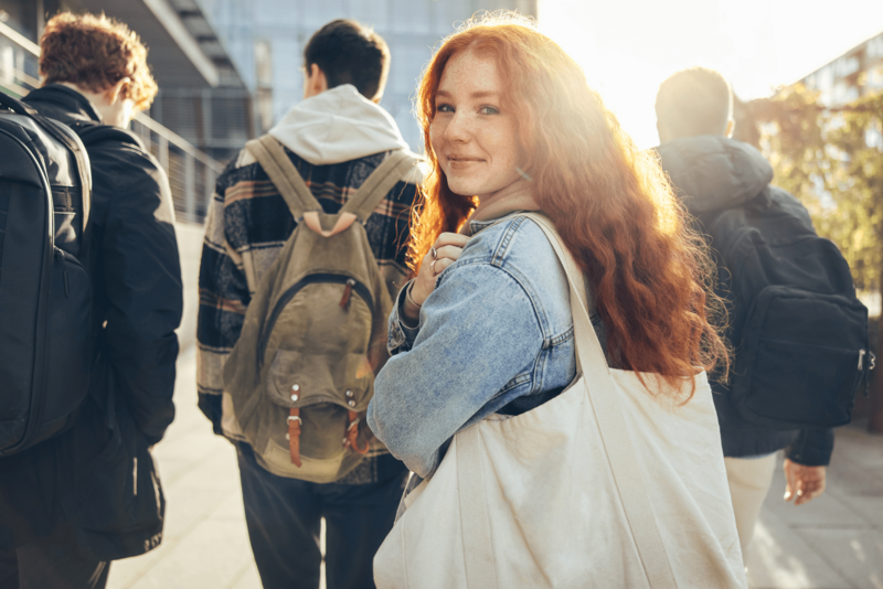 Teenager mit roten Haaren blickt über die Schulter in die Kamera. Im Hintergrund sind drei Jugendliche zu sehen, mit dem Rücken zur Kamera.