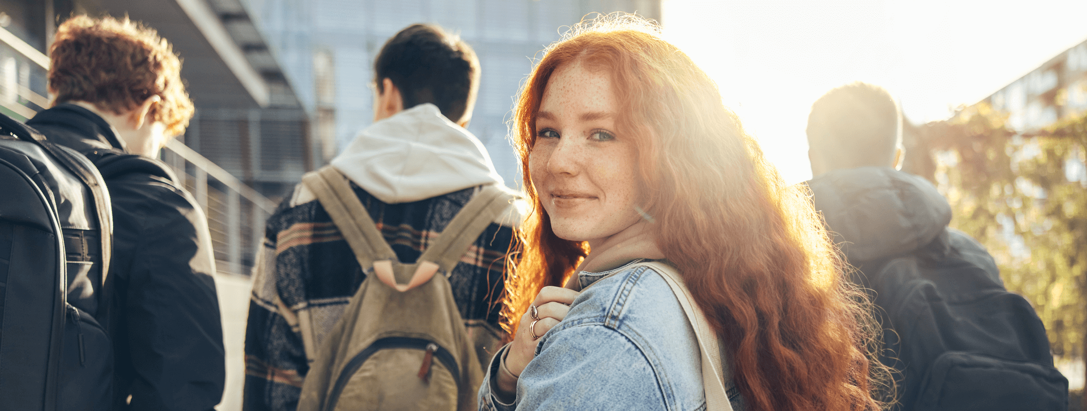 Teenager mit roten Haaren blickt über die Schulter in die Kamera. Im Hintergrund sind drei Jugendliche zu sehen, mit dem Rücken zur Kamera.