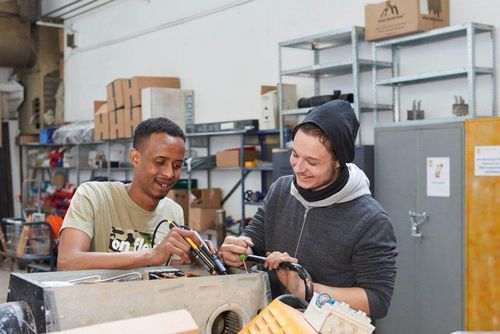 wei Personen arbeiten gemeinsam an einem technischen Gerät in einer Werkstatt. Im Hintergrund sind Regale mit Werkzeugen und Kartons.
