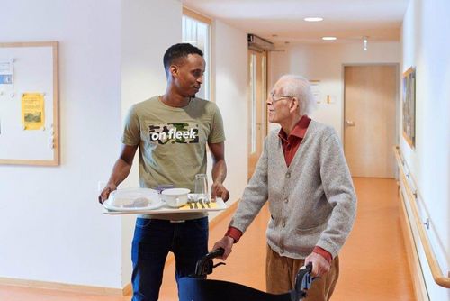 Ein junger Mann trägt ein Tablett mit Geschirr und geht neben einem älteren Mann mit Rollator durch einen hellen Flur im Pflegeheim.
