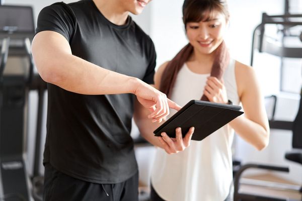 Ein Trainer erklärt einer Kundin ihren Trainingsplan. Er hält ein Tablet in der Hand.