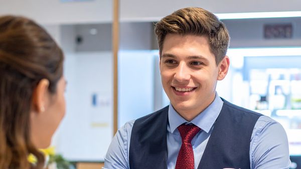 Ein Bankkaufmann Azubi am Schalter im Gespräch mit einer Kundin.