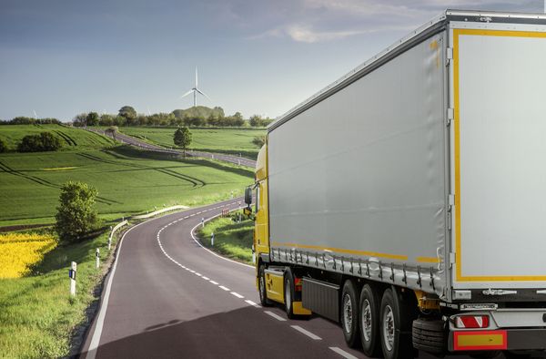 Ein graugelber LKW fährt auf einer kurvigen Landstraße durch eine ländliche Landschaft.