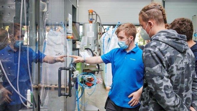 sprungbrett hoponhopoff in schwabmünchen - praktikum bei dittrich Der Auszubildende zeigt den Praktikanten das Bedienpanel der Spritzgussmaschine in einer Fabrikhalle.