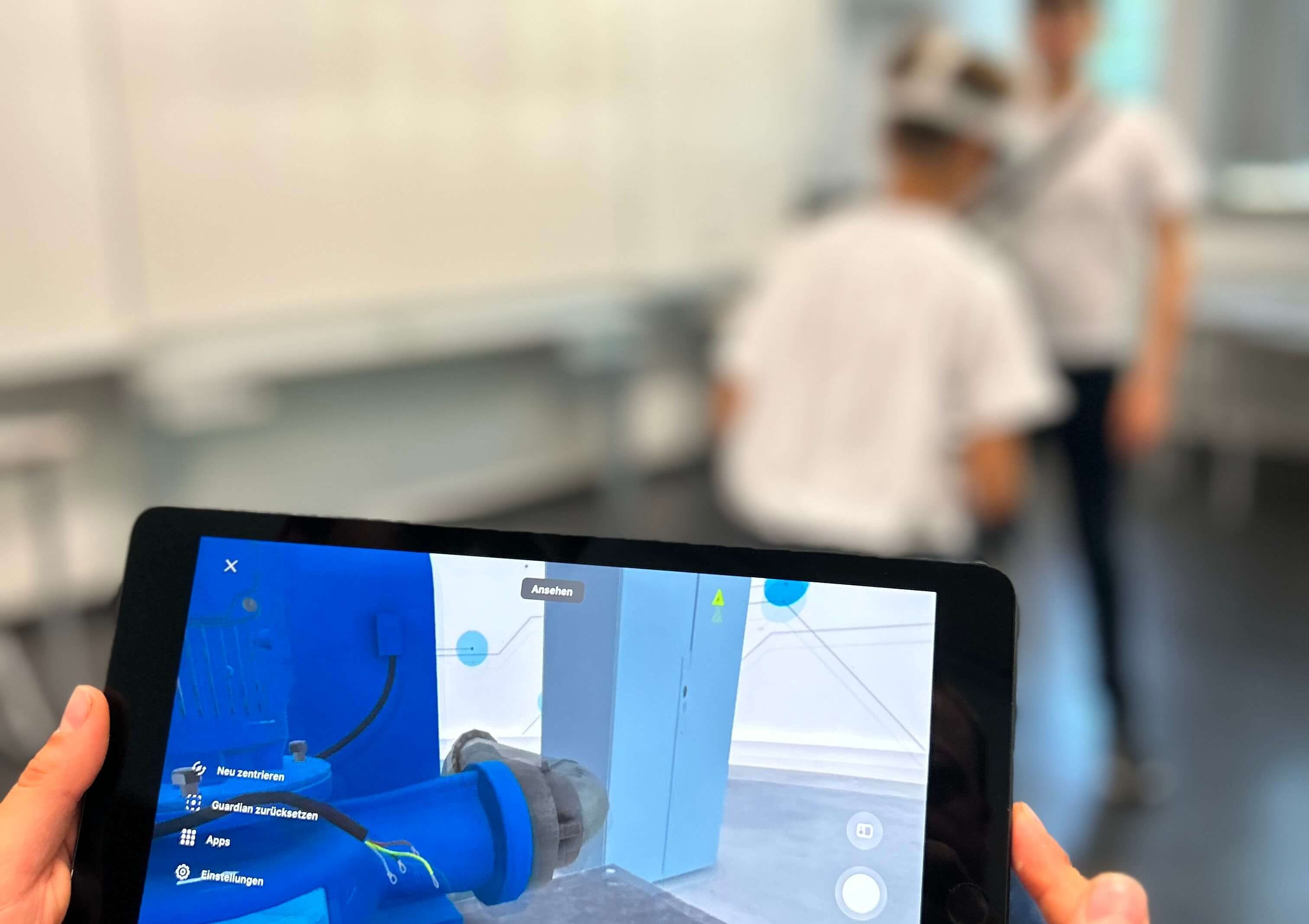 Zwei Schüler im Klassenzimmer spielen die VWE. Im Vordergrund ist ein Tablet abgebildet, welches das Bild aus der VR-Anwendung anzeigt.