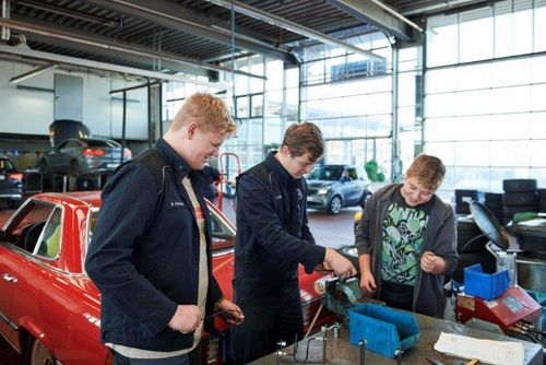 In einer Autowerkstatt stehen drei junge Männer an einer Werkbank. Der Mittlere erklärt den anderen beiden etwas anhand eines Stücks Draht.