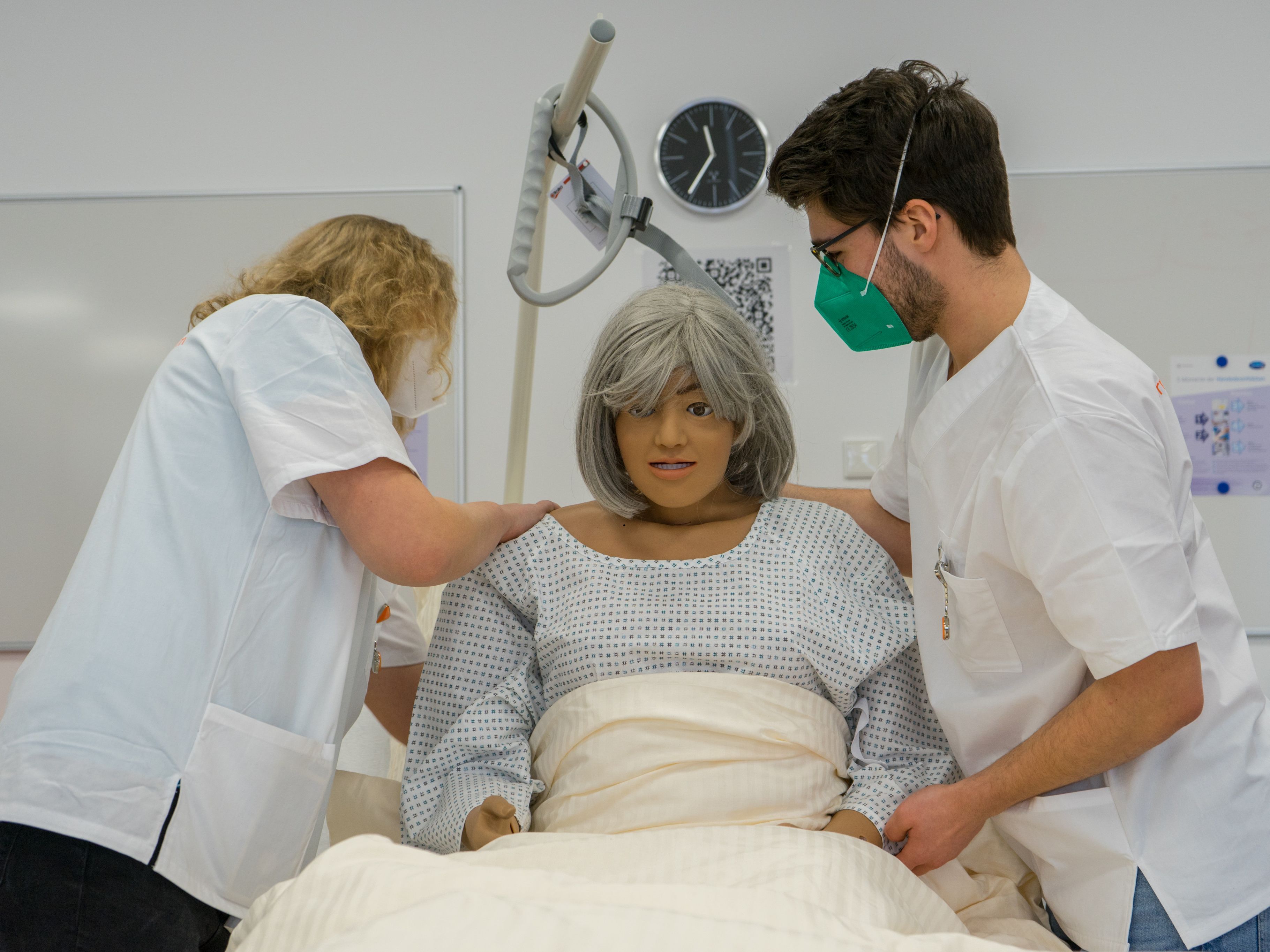 Zwei Studierende üben den richtigen Umgang mit Patient*innen an einer Simulationspuppe, die in einem Krankenhausbett liegt.