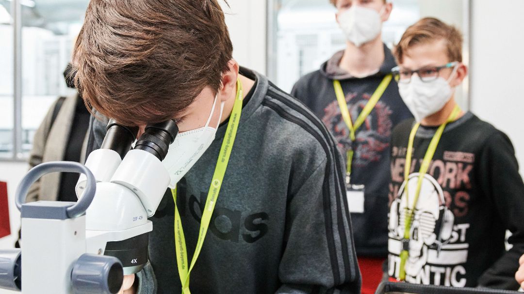 Der Praktikant blickt durch ein Mikroskop, zwei andere Praktikanten sehen ihm zu. alle tragen grüne sprungbrett-Schlüsselbänder