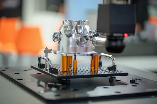 Ein silbernes Metallteil mit Öffnungen und Anschlüssen auf einer schwarzen Plattform mit orangefarbenen Stützen.