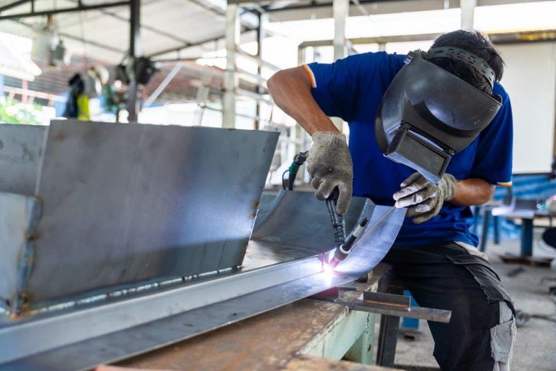 Person mit Schweißhelm und Handschuhen beim Schweißen von Metallteilen – Funken fliegen in industrieller Werkstattumgebung