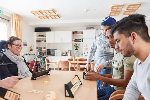 Vier Personen spielen ein Brettspiel an einem Tisch in einem hellen Raum mit Regalen und Schränken im Pflegeheim