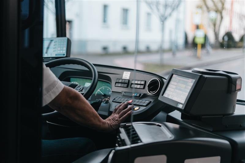 Das Innere einer Busfahrerkabine. Der Fokus liegt auf der Hand des Fahrers, die das Armaturenbrett mit verschiedenen Knöpfen und Schaltern bedient.