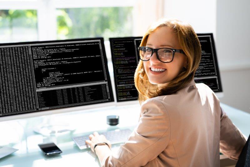 Eine rothaarige Programmiererin sitzt vor zwei Bildschirmen voller Code, sie lacht in die Kamera