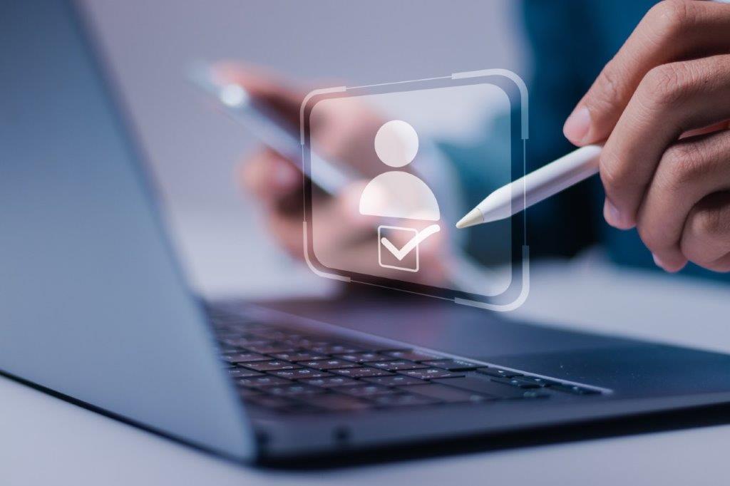 Person interagiert mit Benutzerprofil-Icon auf Laptop per Stylus, Smartphone in der Hand – Symbolbild für digitale Identitätsprüfung oder Kontoverwaltung.