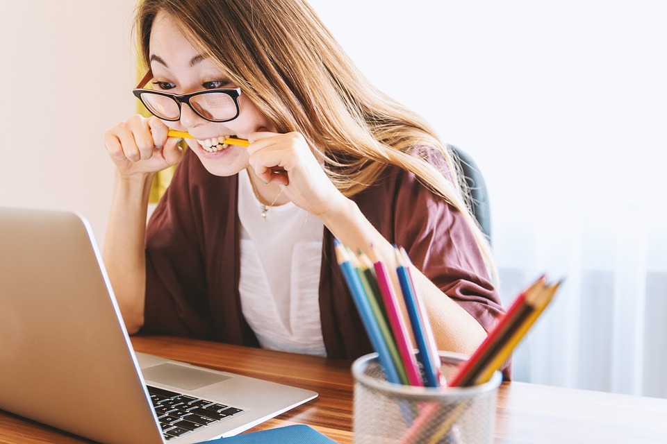 Junge Frau mit Brille sitzt vor einem Laptop und beißt auf einen Bleistift