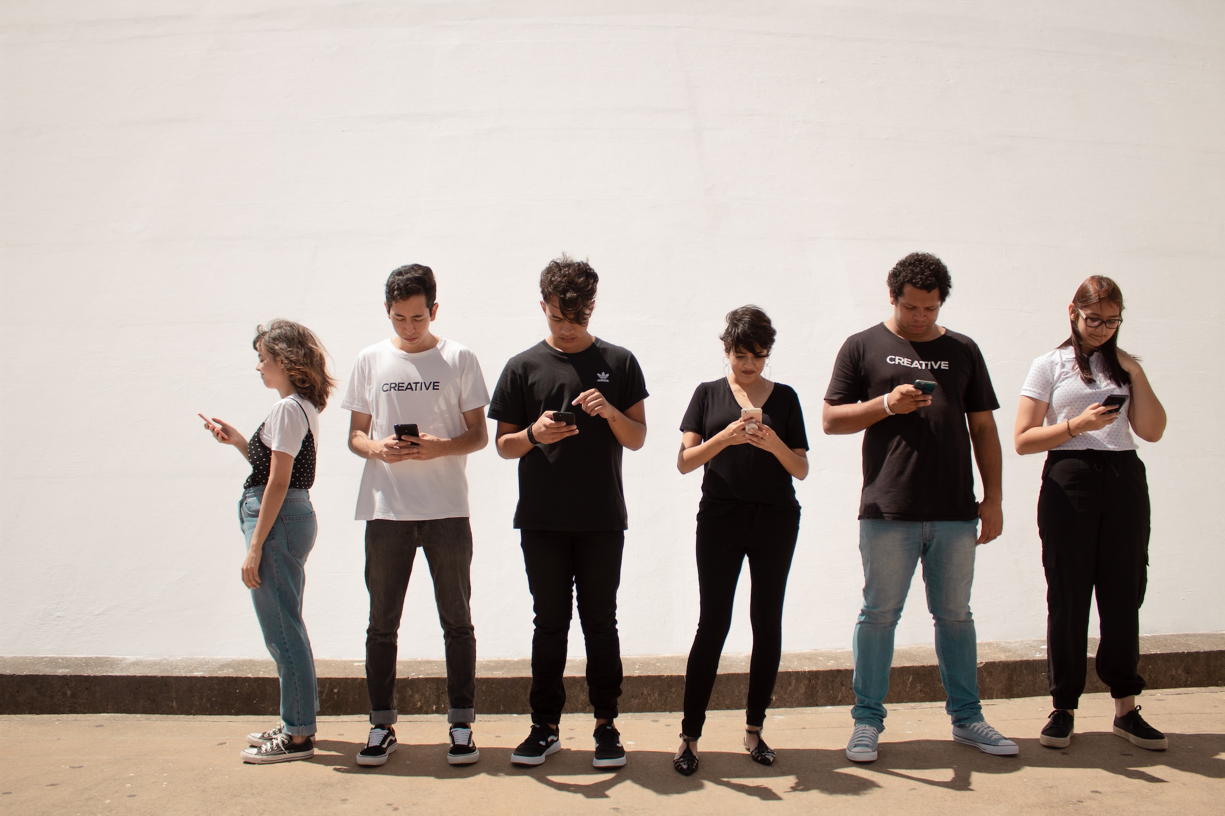 Sechs Teenager stehen vor einer weißen Wand, alle schauen auf ihr Smartphone.