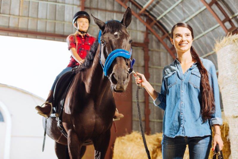 Eine Frau führt ein braunes Pferd am Halfter durch einen Stall. Auf dem Pferd sitzt ein ca 8-jähriges Mädchen. Beide lachen.