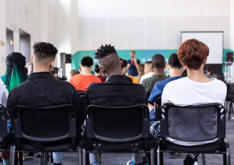 Drei Schüler sitzen in einer Reihe in einem Klassenzimmer und blicken auf ein Whiteboard. Man sieht ihre Rückseite und ihre Stühle. 