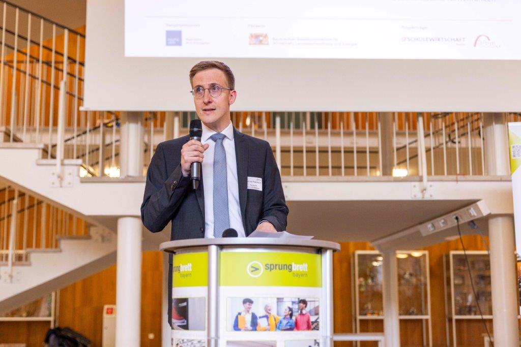 bayme vbm Vertreter am Rednerpult mit Mikrofon beim sprungbrett SPEED DATING – im Hintergrund Treppe und große Leinwand mit Logos und Text.