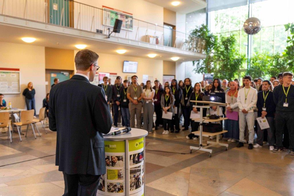 bayme vbm Vertreter spricht am Rednerpult beim sprungbrett SPEED DATING – Publikum steht in der Aula und hört zu.