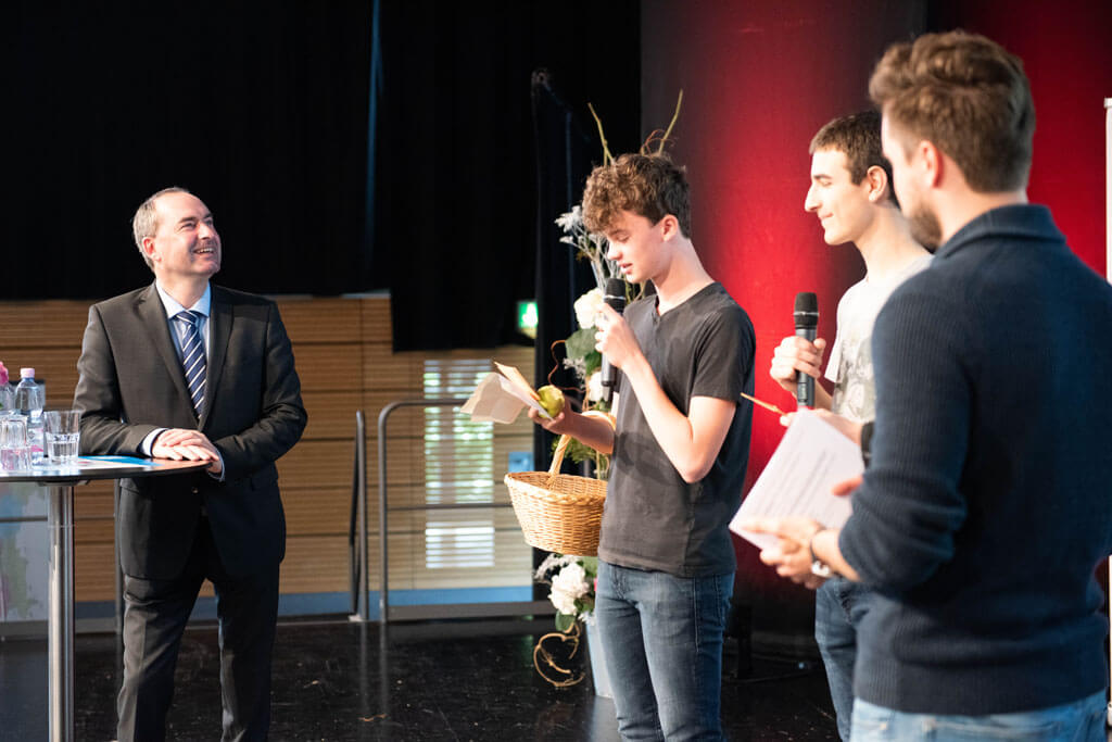 Drei Schüler stellen Wirtschaftsminister Aiwanger auf der Bühne in einem Kurzportrait vor.