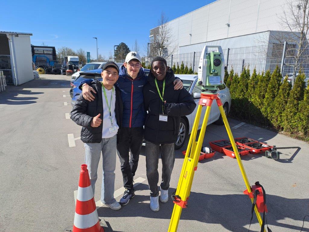 sprungbrett hop-on hop-off Garching 2 Drei Praktikanten stehen im Freien zusammen und posieren für ein Foto mit den Armen umeinander. Im Vordergrund befinden sich ein Verkehrskegel und ein auf einem Stativ montiertes Vermessungsgerät. Im Hintergrund sind geparkte Autos und einige Gebäude zu sehen. Der Himmel ist klar und blau.