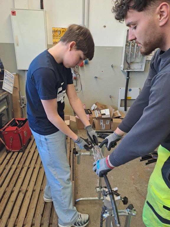 sprungbrett hop-on hop-off Garching 1 Zwei Schüler arbeiten gemeinsam an einem Werkstück in einer Werkstatt. Sie tragen Handschuhe und hantieren mit Metallteilen, möglicherweise beim Zusammenbau oder der Reparatur von Geräten. Um sie herum befinden sich verschiedene Werkzeuge und Kisten.