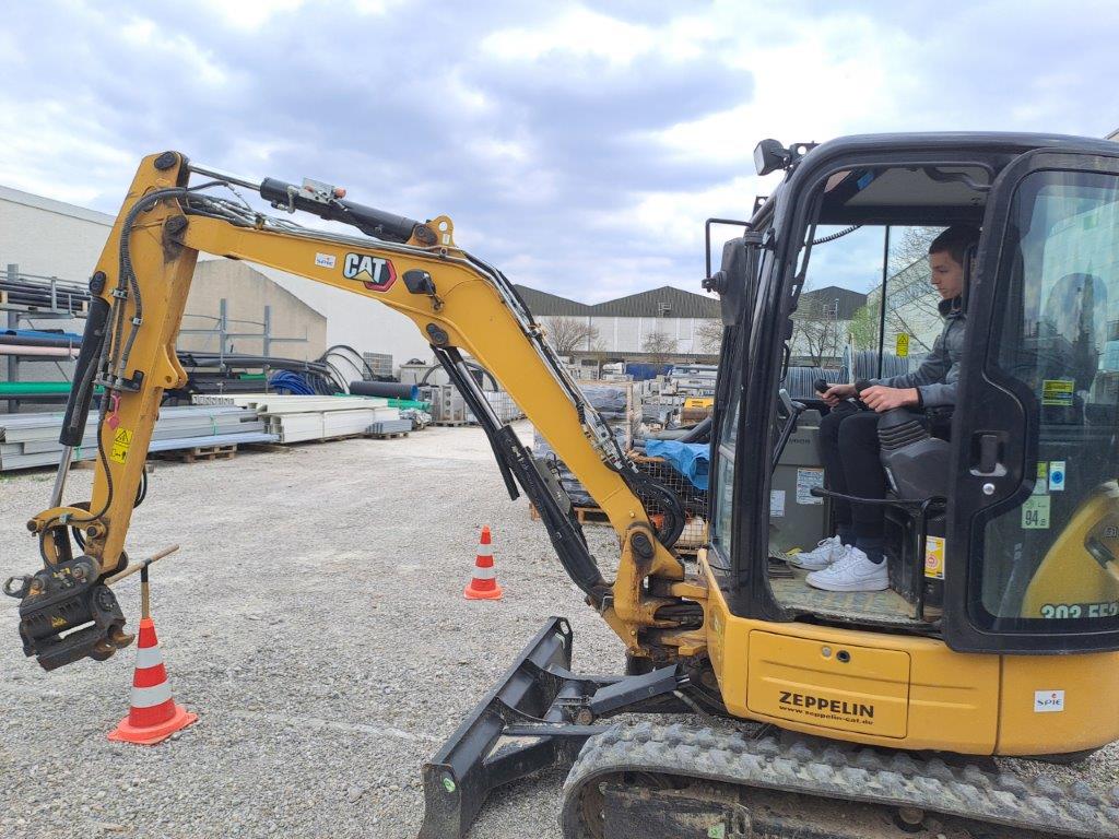 Ein gelber CAT Minibagger mit dem Markennamen 'ZEPPELIN' an der Seite steht auf einer Kiesfläche. Vor dem Bagger sind zwei orange-weiße Verkehrskegel platziert. Im Hintergrund sind verschiedene Baumaterialien wie Metallrohre und -träger sowie Gebäude und andere Geräte zu sehen. Ein Praktikant sitzt in der Kabine des Baggers.