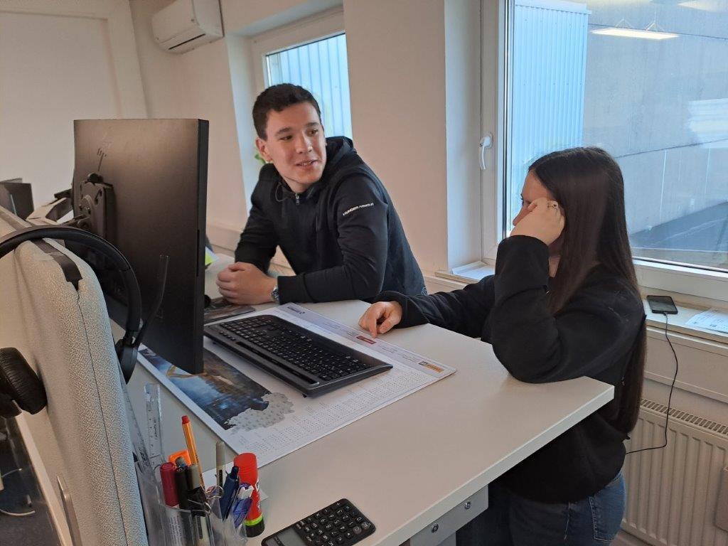 Hop-on hop-off Garching 5 Zwei Personen stehen an einem Schreibtisch mit einem Computerbildschirm, einer Tastatur und einer Maus. Auf dem Schreibtisch befinden sich verschiedene Büromaterialien wie Stifte, Marker und ein Taschenrechner. Im Hintergrund sind Fenster zu sehen.