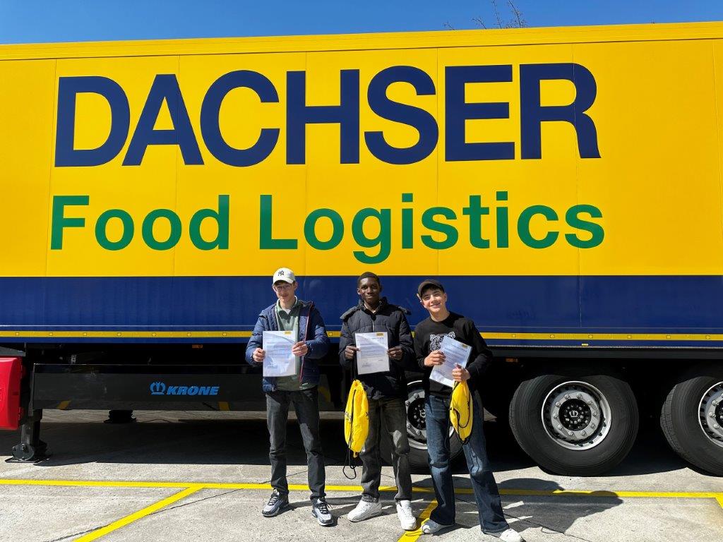 Hop-on hop-off Garching 6 Drei Praktikanten stehen vor einem großen LKW mit der Aufschrift 'DACHSER Food Logistics'. Der LKW ist gelb mit blauer und grüner Schrift. Die Personen halten Papiere und tragen legere Kleidung, jede mit einer gelben Tasche.