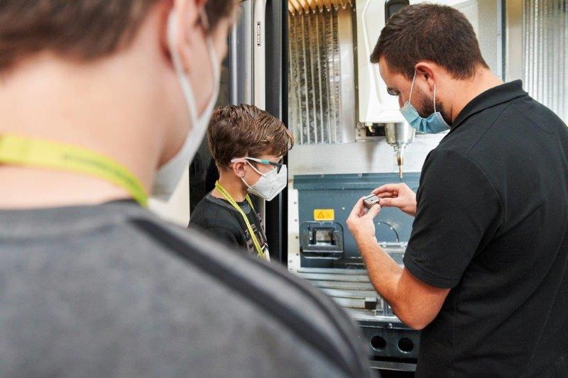 Der Auszubildende erklärt den Praktikanten den Aufbau eines Kleinbauteils. Im Hintergrund eine CNC-Fräse.,