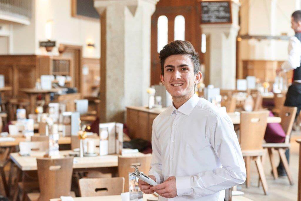 Kellner mit Notizbuch in hellem Restaurant mit Holzmöbeln.