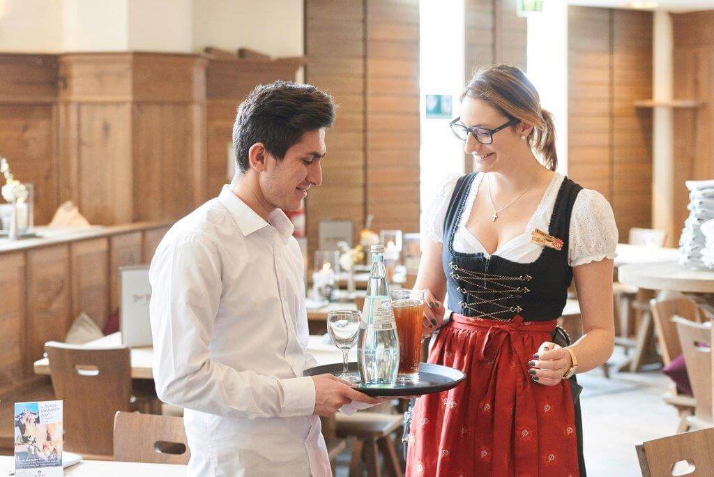 Kellner mit Tablett und Kellnerin in Tracht im Gespräch; Restaurant mit Holzmöbeln im Hintergrund.