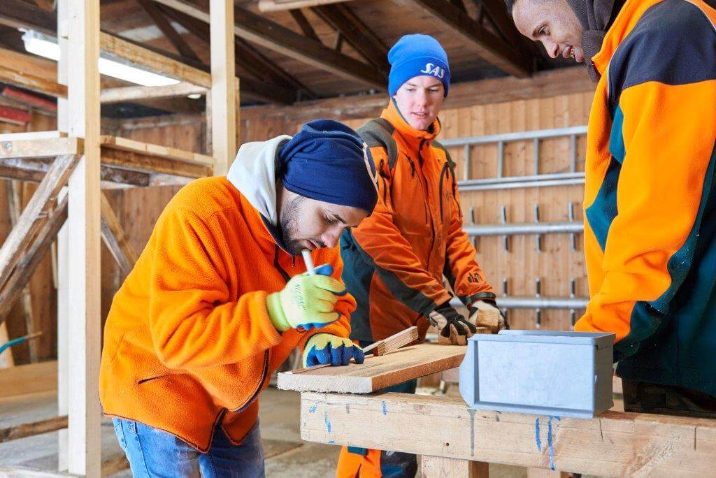 Drei Personen in orangefarbener Arbeitskleidung arbeiten in Werkstatt an einem Holztisch; eine Person sägt Holz.
