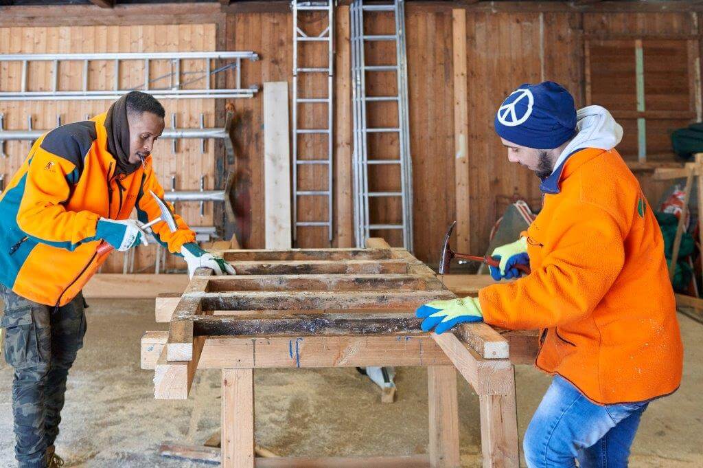 Zwei Männer in orangefarbenen Jacken arbeiten in einer Werkstatt an einem Holzrahmen. Einer hält einen Hammer, der andere eine Zange. Im Hintergrund sind Leitern und Holzregale zu sehen.