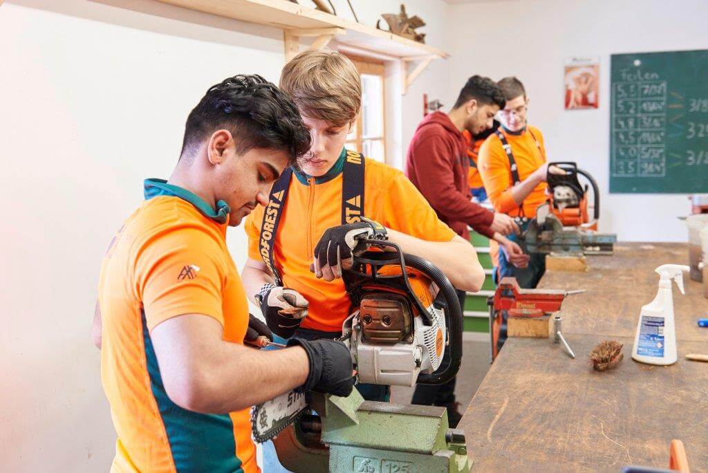 Vier Personen in orange-grünen Uniformen arbeiten in einer Werkstatt an Motorsägen, die in Schraubstöcken eingespannt sind. Im Hintergrund ist eine Tafel mit Text zu sehen.