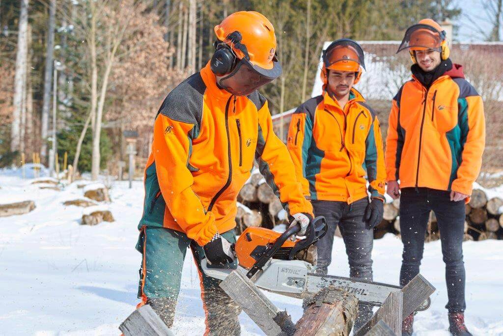 Eine Person in orangefarbener Schutzkleidung sägt mit einer Motorsäge einen Baumstamm im Schnee. Zwei weitere Personen in ähnlicher Kleidung beobachten im Hintergrund.