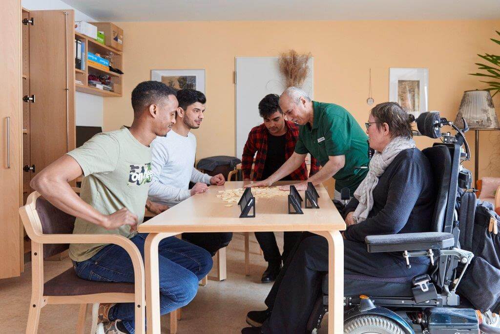 Fünf Personen, darunter eine im Rollstuhl, spielen ein Brettspiel an einem Tisch in einem gemütlich eingerichteten Raum mit Regalen, Bildern und Pflanzen in einem Pflegeheim