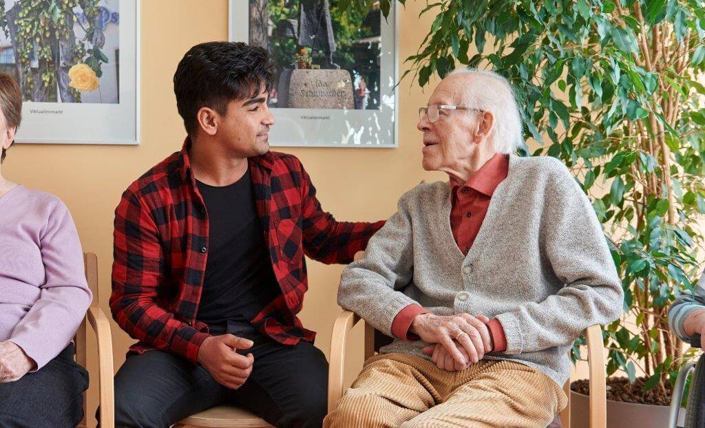  Ein jüngerer Mann in kariertem Hemd sitzt neben einem älteren Mann im grauen Pullover und legt ihm die Hand auf die Schulter. Im Hintergrund sind Pflanzen und Bilder an der Wand.