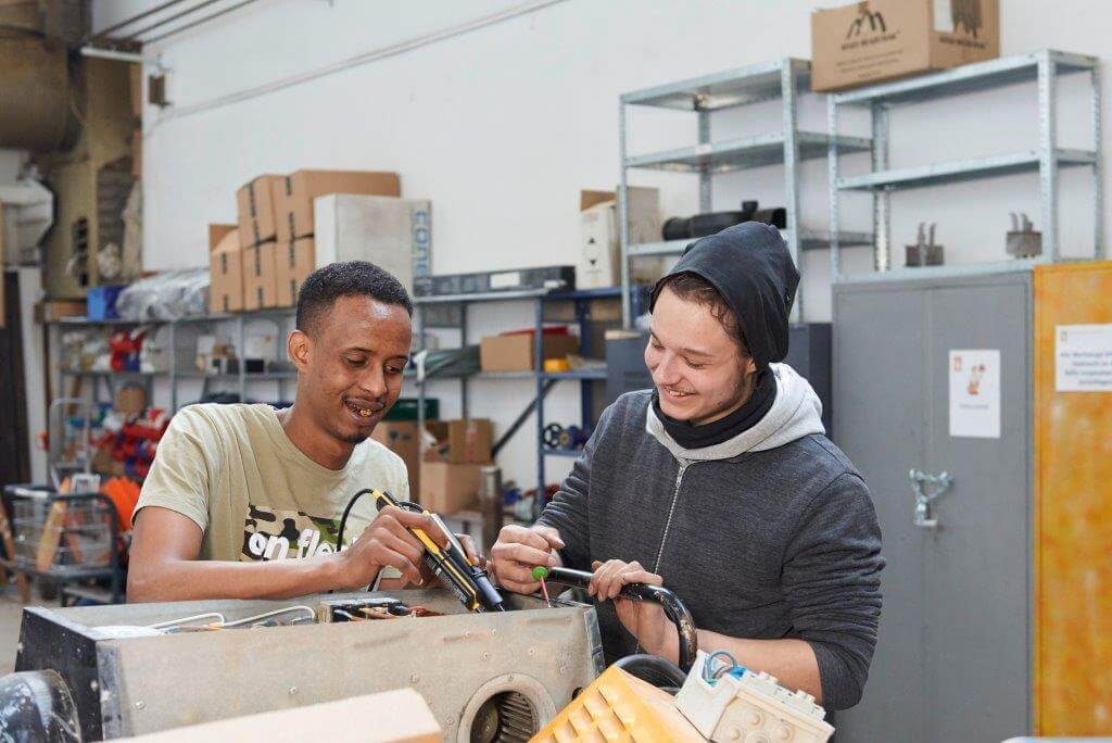 wei Personen arbeiten gemeinsam an einem technischen Gerät in einer Werkstatt. Im Hintergrund sind Regale mit Werkzeugen und Kartons.