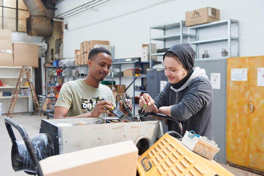 Zwei Personen arbeiten gemeinsam an einem technischen Gerät in einer Werkstatt. Im Hintergrund sind Regale mit Werkzeugen und Kartons.